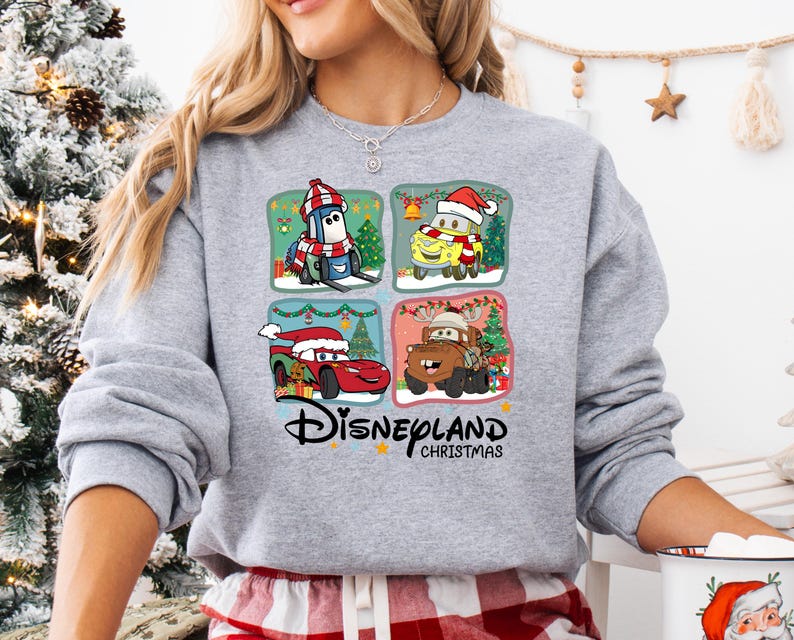 A woman wearing a gray sweatshirt with a Disneyland Christmas-themed design featuring four cartoon cars and a Christmas tree.