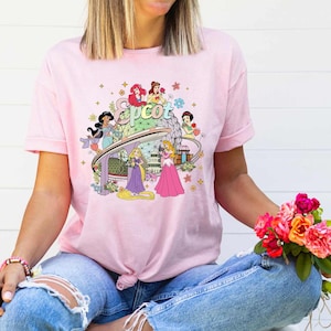 May include: Pink t-shirt with a graphic of Disney princesses and the Epcot logo. The princesses are standing around the Epcot logo and the Spaceship Earth icon.