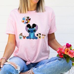 K&ouml;nnte beinhalten: Hellrosa T-Shirt mit einer Cartoon-Figur Stitch, die Mickey Mouse Ohren tr&auml;gt und Luftballons h&auml;lt. Das Shirt hat auch Bilder von Eis und anderen Leckereien. Das T-Shirt ist ein l&auml;ssiger Stil.
