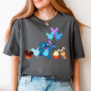 A woman wearing a gray t-shirt with a graphic design featuring a cartoon character surrounded by various Disney-themed items such as Mickey Mouse, cotton candy, and ice cream cones.