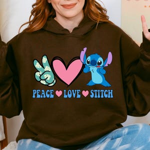 A woman wearing a brown hoodie with a graphic design featuring a peace sign and a heart, along with the text "PEACE LOVE STITCH".
