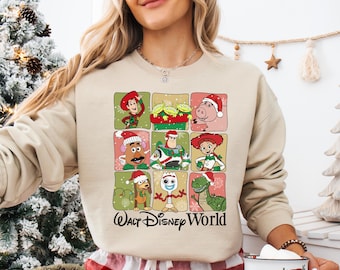 Puede incluir: Sudadera beige con un diseño festivo con personajes de Toy Story con gorros navideños. El diseño de cuadrícula incluye a Woody, Buzz Lightyear y otros, con el texto "Walt Disney World" en letra cursiva.