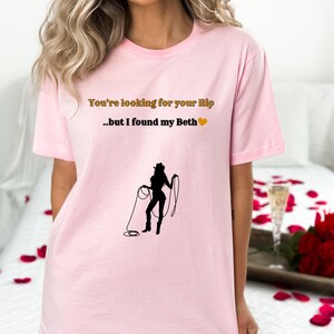 May include: A light pink t-shirt with the text "You're looking for your Rip...but I found my Beth" in gold and a black silhouette of a cowgirl holding a lasso. The shirt is a short-sleeved crew neck.