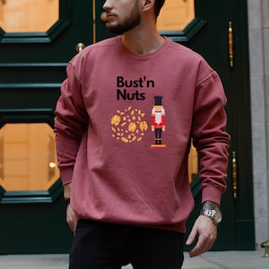 May include: A maroon sweatshirt with the text "Bust'n Nuts" and a cartoon nutcracker holding a bag of nuts.