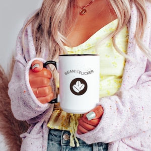 May include: White coffee mug with a black rim and handle, featuring the text "Beans of Flicker" and a brown coffee bean logo. The mug is held by a person wearing a light purple sweater and a yellow floral top.