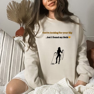 May include: A cream-colored sweatshirt with the text "You're looking for your Rip...but I found my Beth" and a silhouette of a woman roping. The text is in a gold color and the heart is orange.