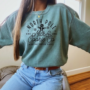 May include: A teal green sweatshirt with a black graphic design that says "North Pole Milk & Cookie Co. Est. 1984 Baking Fresh Daily" with a gingerbread man illustration.
