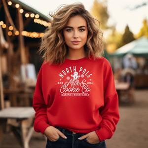 May include: Red sweatshirt with a white graphic design that says "North Pole Milk & Cookie Co. Est. 1984 Baking Fresh Daily" with a gingerbread man illustration.