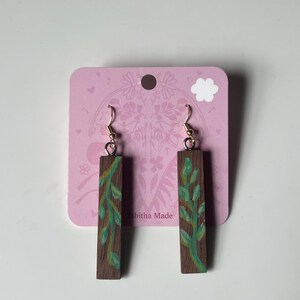 May include: Wooden earrings with a rectangular shape and a painted green vine design. The earrings hang from gold-colored hooks. The earrings are displayed on a pink card with the text "Tabitha Made".