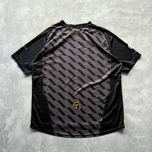 May include: Black and gray Real Madrid soccer jersey with a geometric pattern on the back. The jersey has gold accents on the shoulders and around the collar. The words "REAL MADRID" are printed across the upper back.