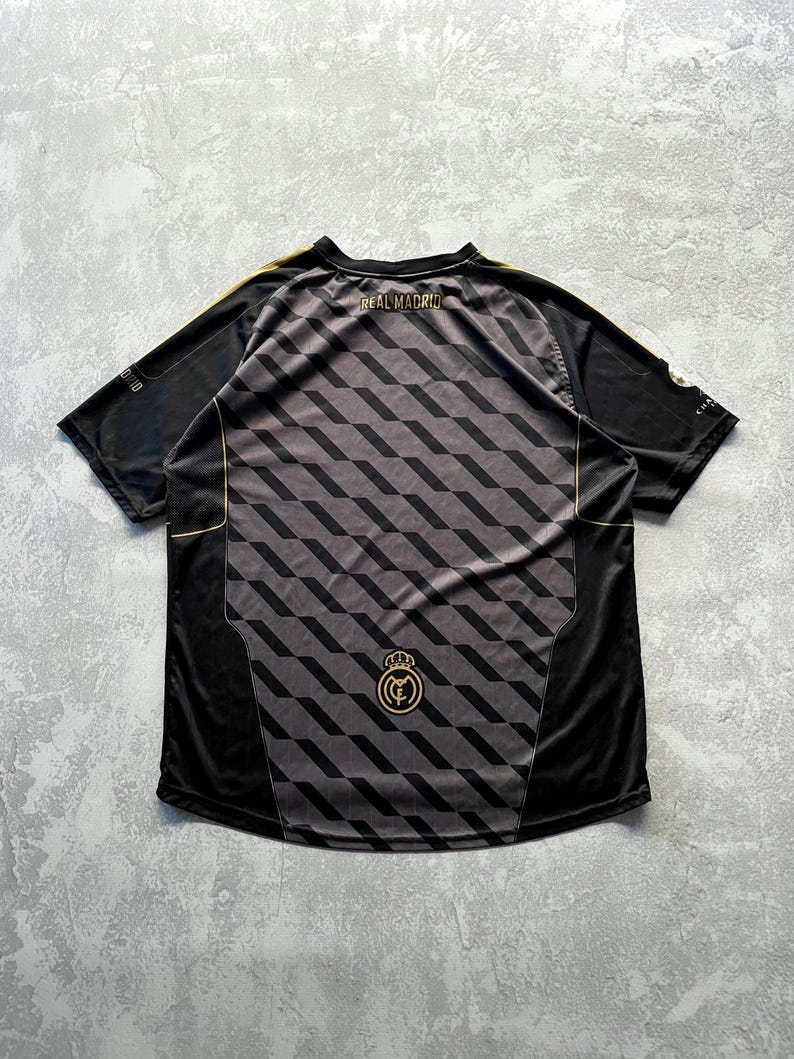May include: Black and gray Real Madrid soccer jersey with a geometric pattern on the back. The jersey has gold accents on the shoulders and around the collar. The words "REAL MADRID" are printed across the upper back.