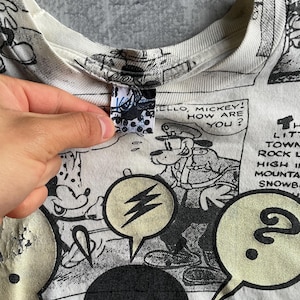 May include: White t-shirt featuring a black and white comic strip design with Mickey Mouse and other characters. Speech bubbles with text and a lightning bolt graphic are visible. The shirt's collar and tag are also visible.