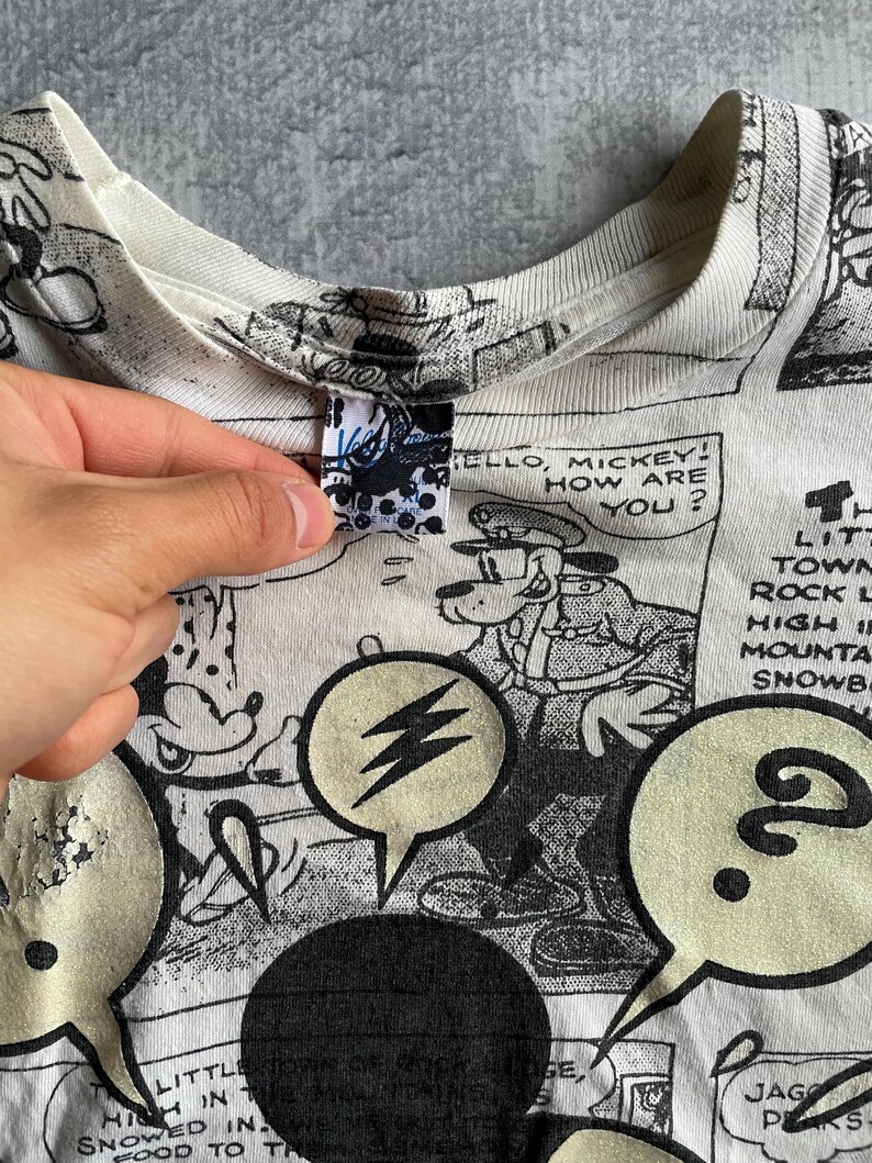 May include: White t-shirt featuring a black and white comic strip design with Mickey Mouse and other characters. Speech bubbles with text and a lightning bolt graphic are visible. The shirt's collar and tag are also visible.