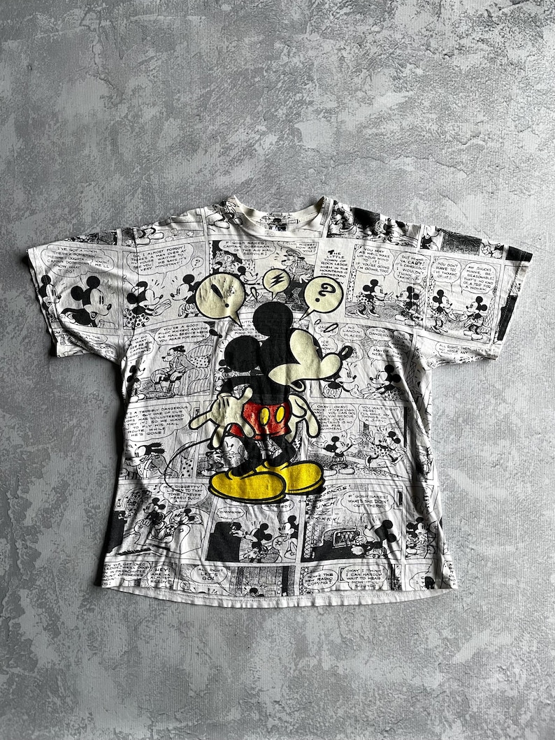 May include: White t-shirt featuring a large Mickey Mouse graphic with a comic strip design. Mickey is in his classic pose, wearing red shorts and yellow shoes. The shirt has a crew neck and short sleeves.