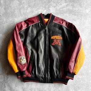 May include: A black leather jacket with maroon and gold accents. The jacket features the word "Gophers" and a large "M" in orange on the chest. A cartoon gopher patch is on the left sleeve. The jacket has a zipper closure and ribbed cuffs.