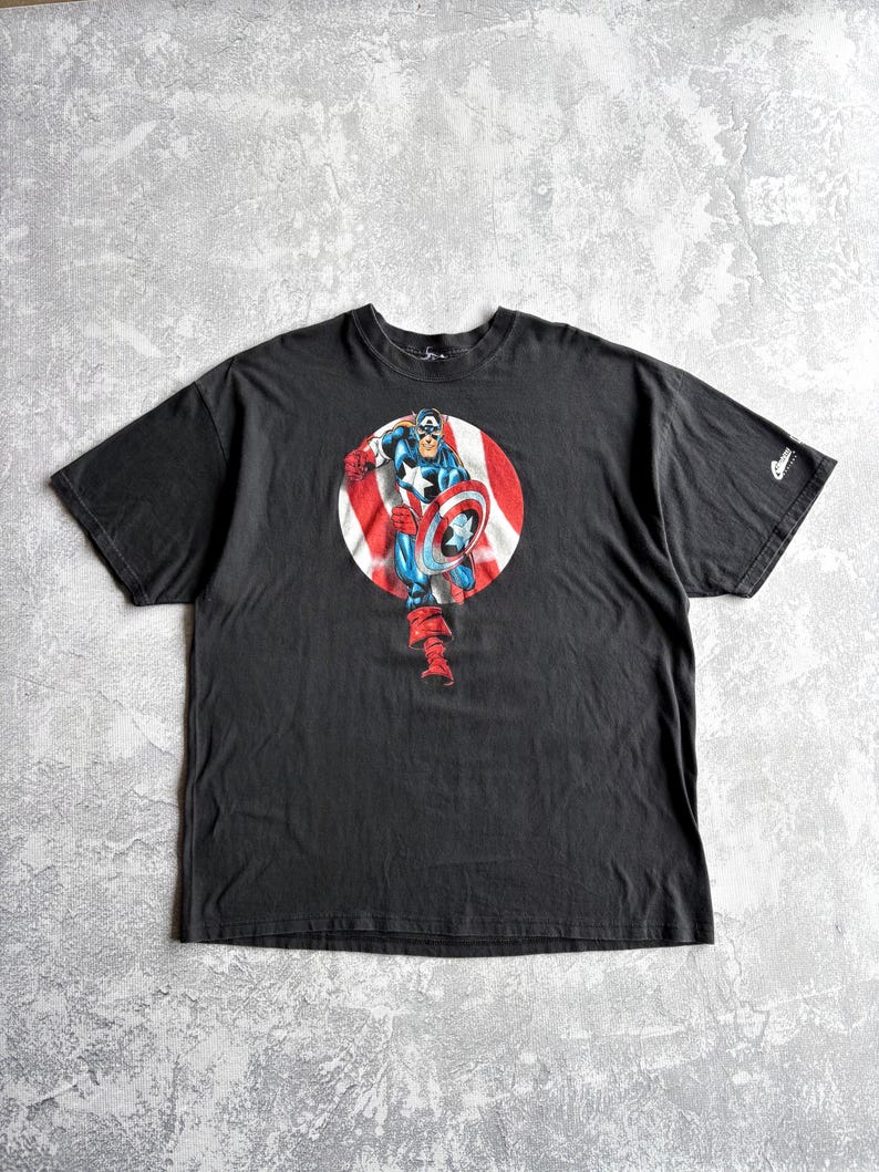 May include: Black t-shirt featuring a Captain America graphic. The graphic shows Captain America in a red, white, and blue circle, holding his shield. The shirt has short sleeves and a crew neck.