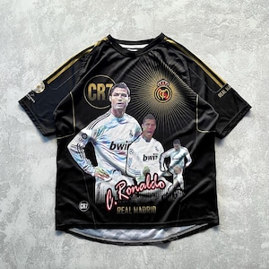 May include: Black Real Madrid soccer jersey featuring images of Cristiano Ronaldo. The jersey has gold accents on the shoulders and the text "C. Ronaldo" in red script. The Real Madrid crest is also visible.