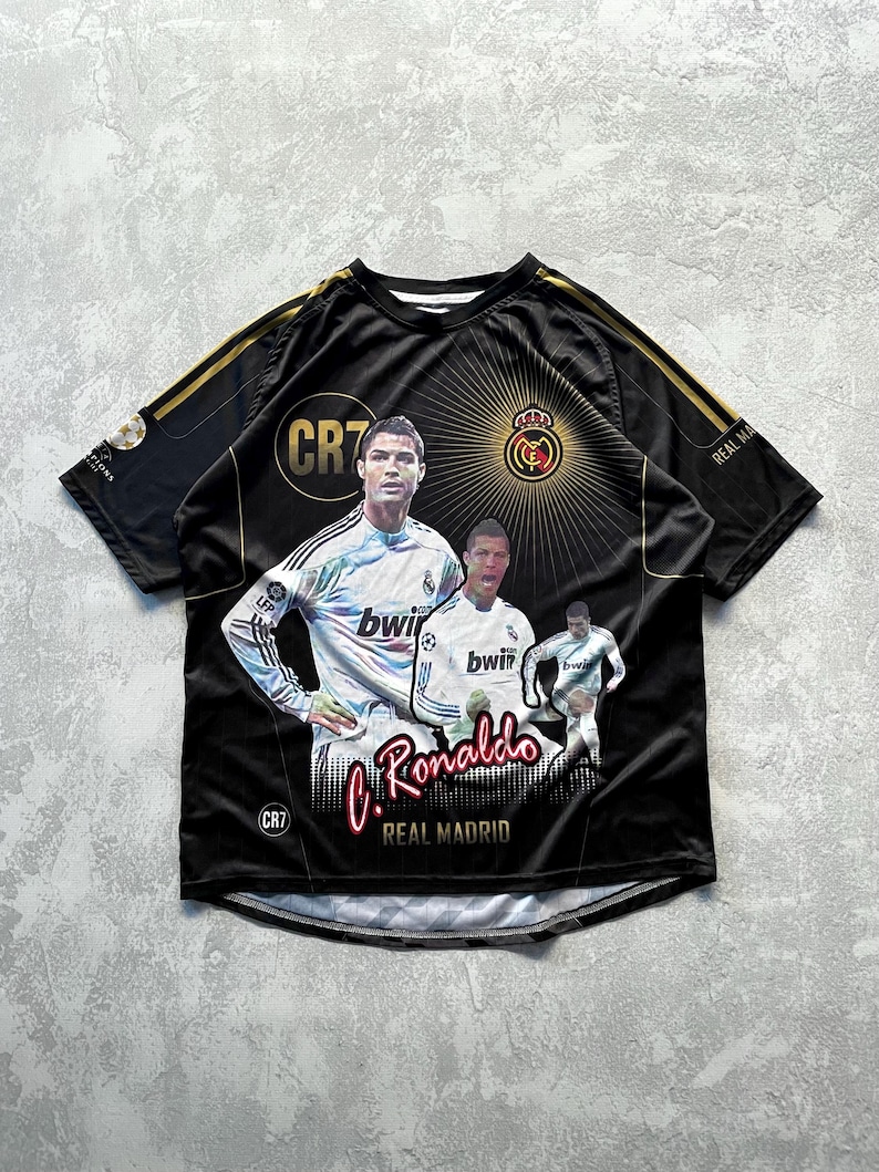May include: Black Real Madrid soccer jersey featuring images of Cristiano Ronaldo. The jersey has gold accents on the shoulders and the text "C. Ronaldo" in red script. The Real Madrid crest is also visible.