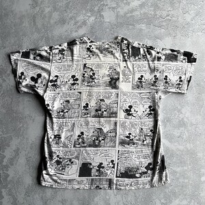 May include: Black and white t-shirt featuring a comic strip design with Mickey Mouse and other characters. The shirt has short sleeves and a crew neck. The comic strip panels are arranged across the entire shirt.