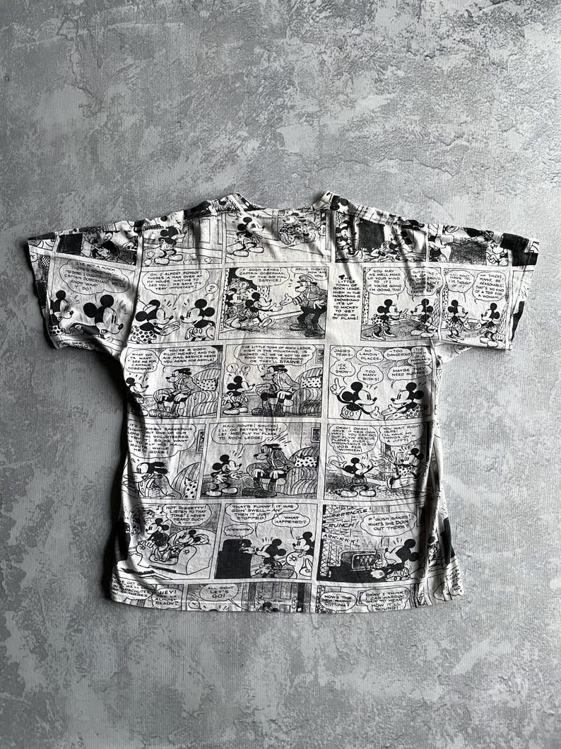 May include: Black and white t-shirt featuring a comic strip design with Mickey Mouse and other characters. The shirt has short sleeves and a crew neck. The comic strip panels are arranged across the entire shirt.