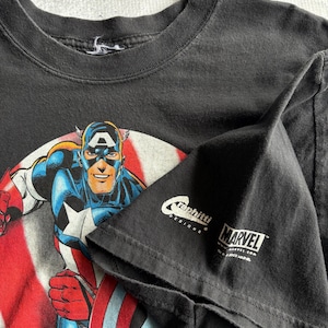 May include: Black t-shirt featuring Captain America in a dynamic pose, set against a backdrop of the American flag. The shirt includes the Marvel logo and the text "Graphitti Designs."