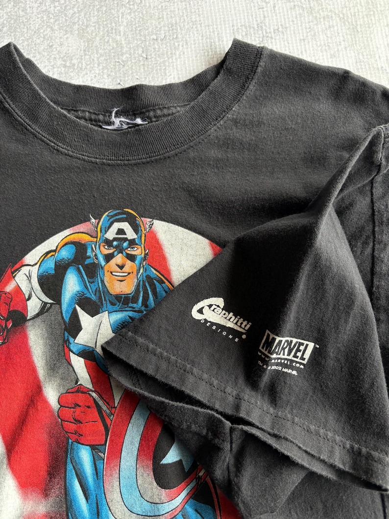 May include: Black t-shirt featuring Captain America in a dynamic pose, set against a backdrop of the American flag. The shirt includes the Marvel logo and the text "Graphitti Designs."
