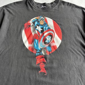 May include: Dark gray t-shirt featuring a graphic of Captain America in a running pose, holding his shield. The character is set against a red and white striped circle. The shirt is made of a soft material.