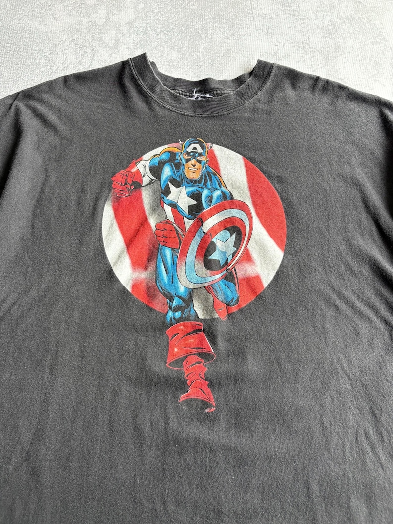 May include: Dark gray t-shirt featuring a graphic of Captain America in a running pose, holding his shield. The character is set against a red and white striped circle. The shirt is made of a soft material.