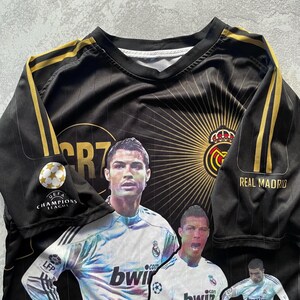May include: Black and gold Real Madrid soccer jersey featuring images of Cristiano Ronaldo. The jersey has gold stripes on the sleeves and the UEFA Champions League logo. The Real Madrid crest is visible.