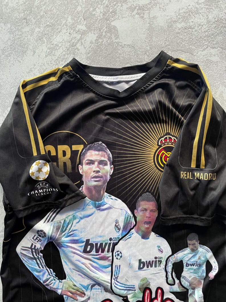 May include: Black and gold Real Madrid soccer jersey featuring images of Cristiano Ronaldo. The jersey has gold stripes on the sleeves and the UEFA Champions League logo. The Real Madrid crest is visible.