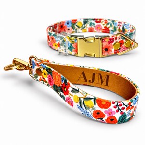May include: A pet collar and a matching wristlet strap featuring a vibrant floral pattern with red, orange, yellow, and blue flowers on a white background. Both items include gold-toned metal hardware, and the strap is engraved with the letters AJM.