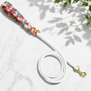 May include: White dog leash with a floral patterned handle featuring red, orange, and blue flowers. The leash has gold-colored hardware and a white rope design. The leash is designed for pets.