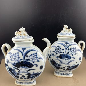 May include: Two antique white and blue ceramic teapots with intricate floral and bird designs. Each teapot has a small handle, a spout, and a lid topped with a small animal figurine. The teapots are displayed on a neutral surface.