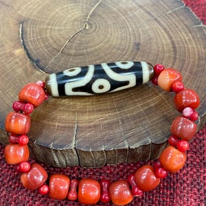 May include: A bracelet with a central black and white patterned bead. The bracelet is made of red coral beads of different shapes and sizes. The bracelet is displayed on a wooden surface. The beads are strung together.