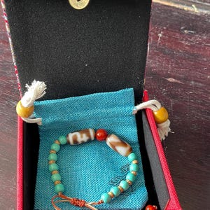 May include: A turquoise beaded bracelet with two patterned beads and a red bead, presented in a red box with a blue pouch. The box has a decorative lid with a gold coin accent.