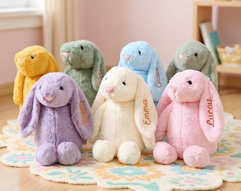 May include: A collection of plush bunny rabbits in various pastel colors, including yellow, green, blue, purple, cream, and pink. Each bunny has long floppy ears and embroidered names. The bunnies are sitting on a floral patterned rug.