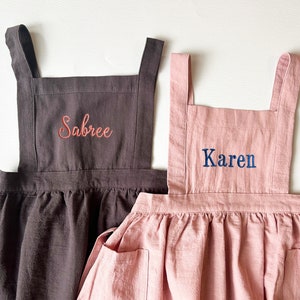 May include: Two personalised linen aprons, one brown with the name "Sabree" embroidered in pink and one pink with the name "Karen" embroidered in blue.