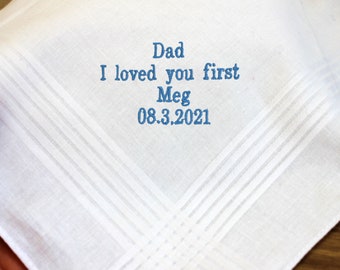 May include: White handkerchief with blue embroidered text. The text reads "Dad I loved you first Meg 08.3.2021". The handkerchief has a subtle striped pattern and is folded.