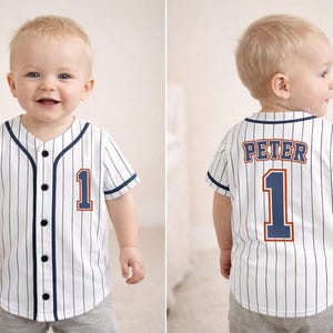 Puede incluir: Un bebé con una camiseta de béisbol blanca con finas rayas azul marino, ribetes y botones. La camiseta tiene el número 1 y el nombre "PETER" en la espalda. El bebé está sonriendo.