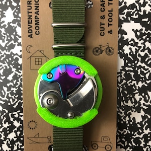 May include: A multi-tool watch with a green nylon strap and a green foam case. The watch face is a circular metal disc with a rainbow iridescent finish. The packaging has the text "Adventure's Companion" and "Cut & Carve & Tool Tips".