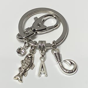 May include: Silver keychain with a fish charm, a letter A charm, a hook charm with the text "Hook me", and a clear rhinestone charm.