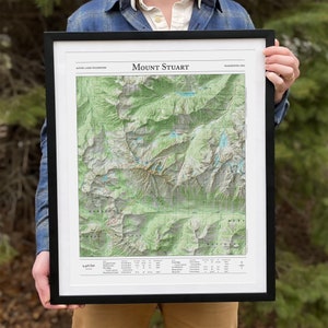 May include: A framed map of Mount Stuart in the Alpine Lakes Wilderness, Washington, USA. The map shows topographic details of the area, including mountains, lakes, and trails. The map is printed on white paper and has a black frame.
