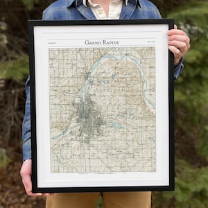May include: A framed vintage map of Grand Rapids, Michigan. The map is printed in black and white with brown and blue accents. The map shows the city's streets, rivers, and other features.
