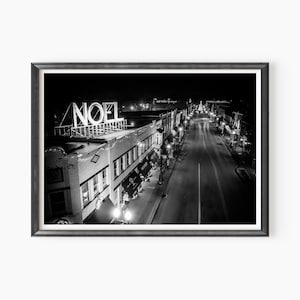 May include: A black and white photograph of a city street at night with a large illuminated sign that reads "NOEL" on top of a building. The street is lined with buildings and streetlights.