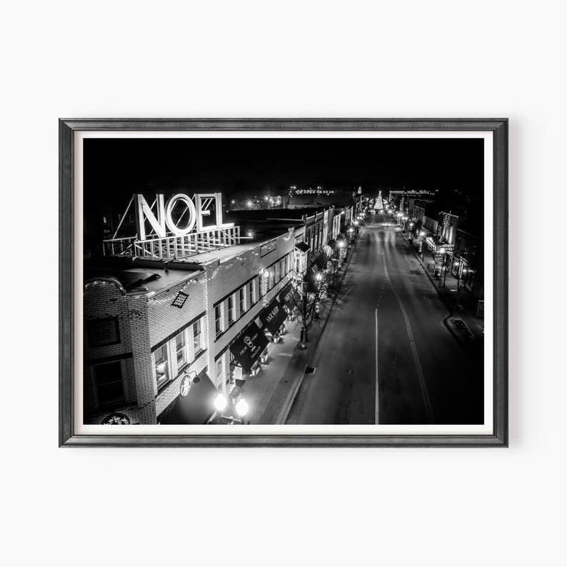May include: A black and white photograph of a city street at night with a large illuminated sign that reads "NOEL" on top of a building. The street is lined with buildings and streetlights.