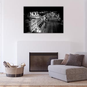 May include: Black and white photograph of a city street at night with a sign that reads "NOEL" above a building. The street is lined with buildings and streetlights.