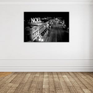 May include: Black and white photograph of a city street at night with a sign that reads "NOEL" above the buildings. The street is lined with buildings and streetlights.