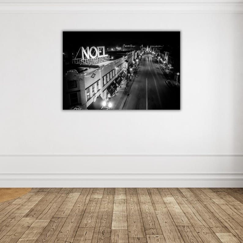 May include: Black and white photograph of a city street at night with a sign that reads "NOEL" above the buildings. The street is lined with buildings and streetlights.