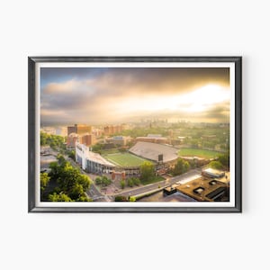May include: An aerial view of a football stadium surrounded by buildings and trees. The stadium is surrounded by a green field and has a white roof. The sky is cloudy and the sun is shining through the clouds.