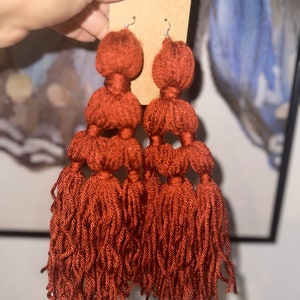 Op de afbeelding: Een paar lange, roestkleurige kwast oorbellen met drie lagen pompoenen. De oorbellen zijn gemaakt van garen en hebben een zilveren haak.
